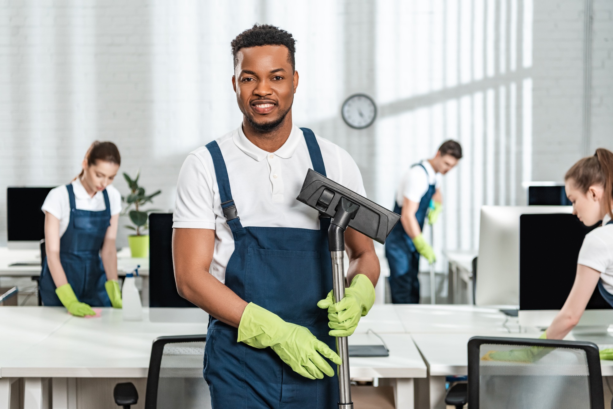 smiling african american cleaner holding vacuum cleaner brush near team of multicultural colleagues.